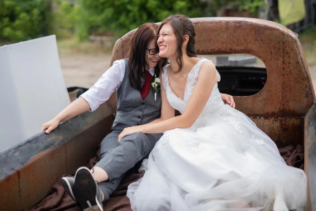 Isabell und Sonya Nubel, Hochzeitsfoto, zwei Frauen auf einem rostigen Auto. Isabell Nubel trägt einen grauen Anzug mit einer roten Krawatte. Sonya Nubel trägt ein langes weißes Brautkleid. Beide sind sehr innig miteinander und lachen. Queeres Brautpaar 